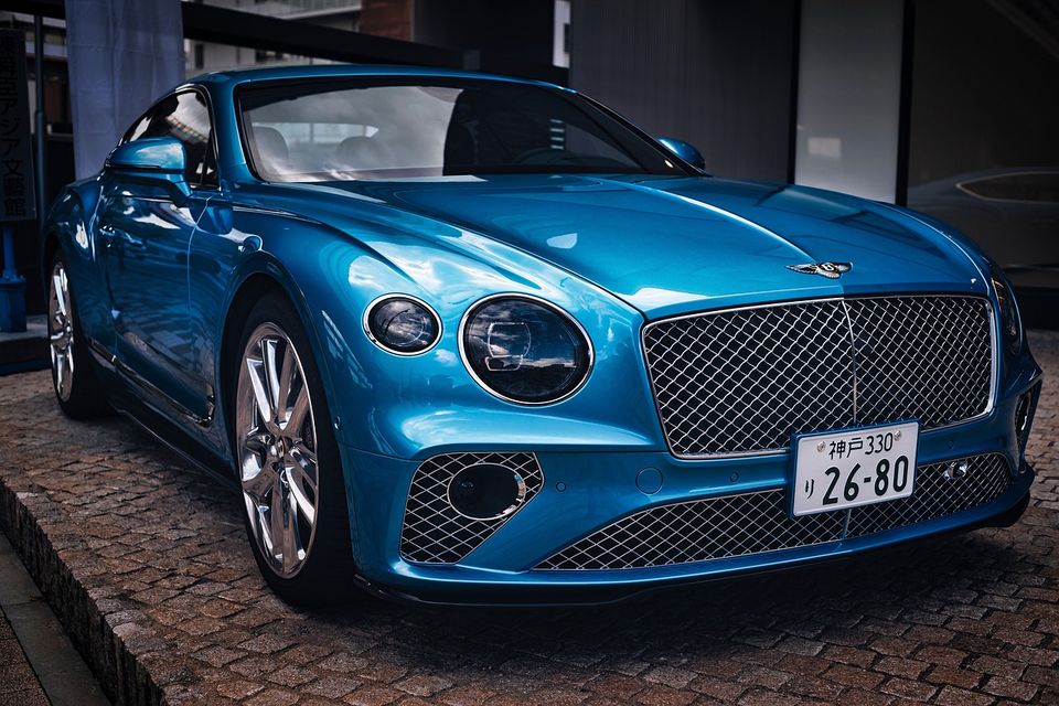 Image of a blue Bentley from the passenger side quarter front view, Parked wheels are straigtht, flourescent lighting