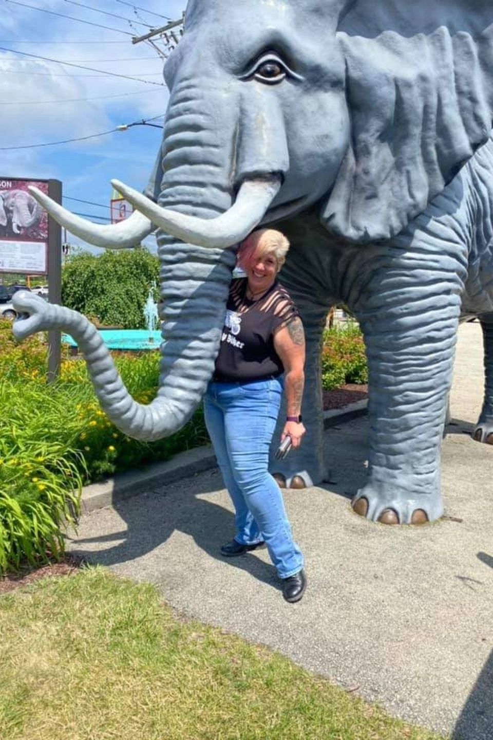 Woman posing beneath a large elephant statue outdoors on a sunny day.