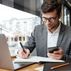 Smiling businessman eyeglasses sitting by table cafe with laptop computer while using smartphone writing something