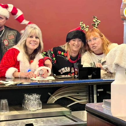 Three women in holiday-themed outfits smiling behind an event registration or service counter.