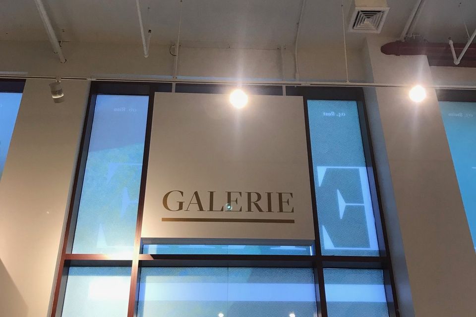 Illuminated panel reading Galerie mounted above tall glass entry doors inside a building lobby