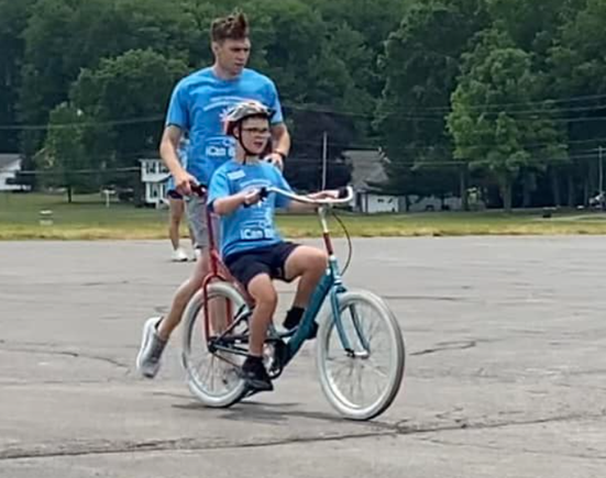 A volunteer jogs beside a participant riding an adaptive bicycle in an outdoor parking lot during DSAV's iCan Shine Bike Camp.