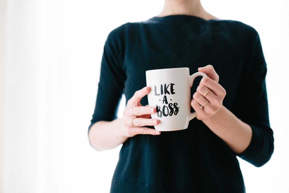 A person wearing a dark shirt holds a white coffee mug with the words 'LIKE A BOSS' printed on it, standing against a bright, light-colored background
