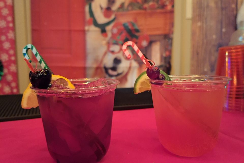 Two colorful cocktails with fruit garnish and candy cane on a red table