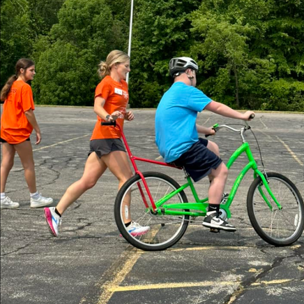 A volunteer coach runs beside a participant riding an adaptive bicycle in an outdoor practice area during DSAV's iCan Shine Bike Camp.