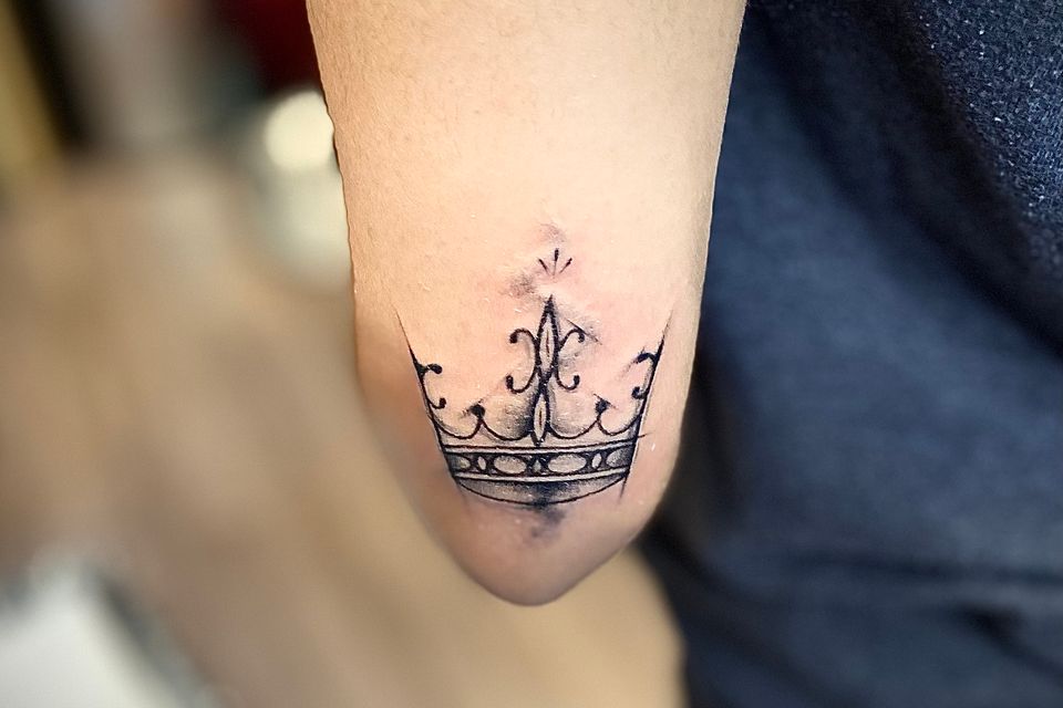 Image of a tattoo near the elbow at the top of the forearm in black and grey of a Kings Crown