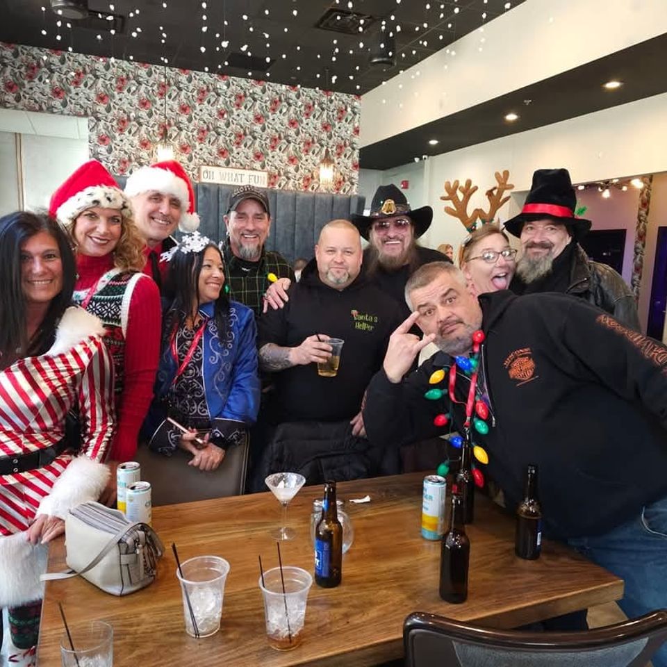 Group of attendees wearing Christmas-themed clothing and accessories gathered around a table at a holiday party indoors.