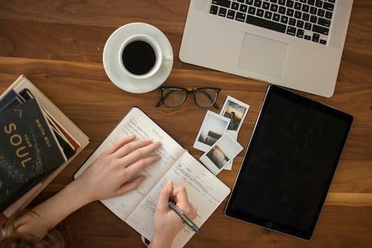Person's hands writing in a journal with glasses, coffee, photos, books and laptop about 