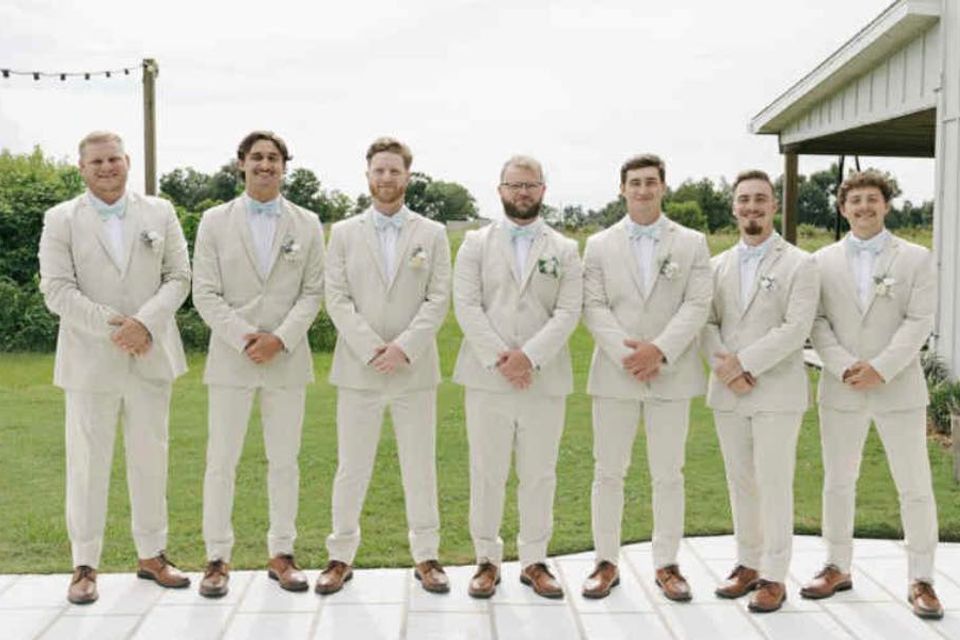 Groomsmen wearing matching light beige suits with white shirts and pastel bow ties, posing outdoors at a wedding.