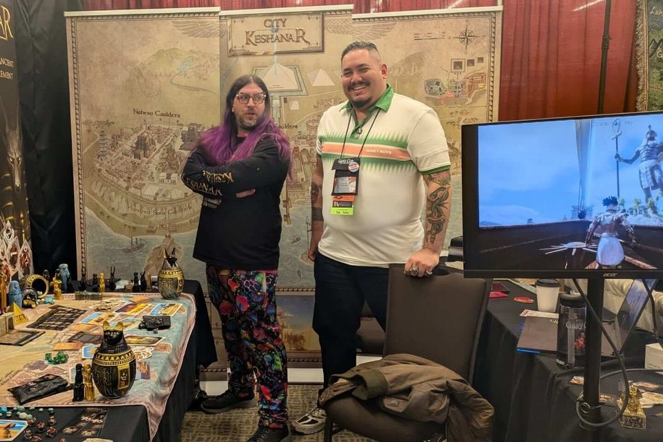 Two RPG creators at a convention booth with a City of Keshanar map backdrop, table display of miniatures and game props, and demo screen
