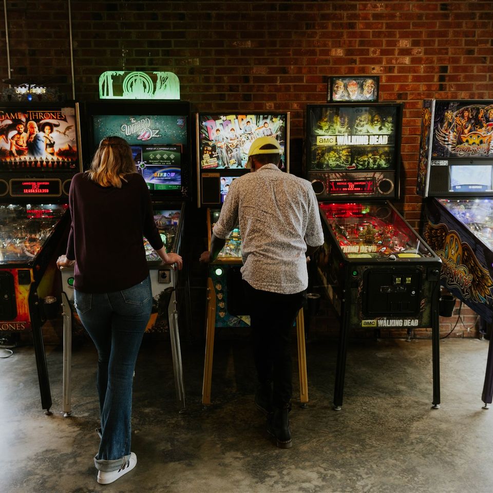 Two people playing two different pinball machines with three more in view