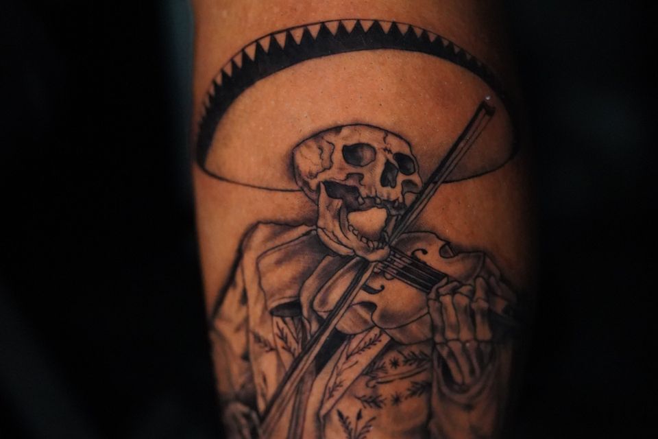 Iamge of a left calf male tattoo of a skeleton wearing a sombrero, dressed in a decorative jacket in black & grey ink