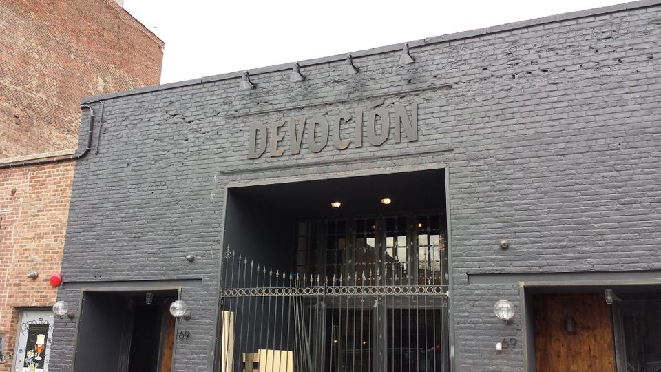 Grey dimensional letters reading Devoción mounted above a gated entrance on a painted dark grey brick facade