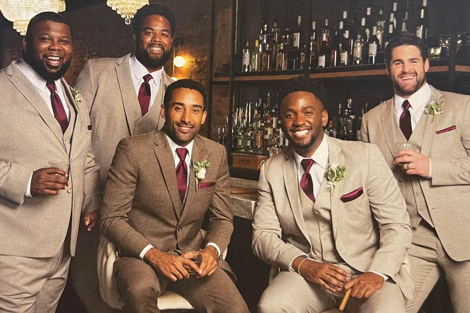 Wedding party wearing coordinated tan suits with burgundy ties and pocket squares from New Orleans Tuxedo Inc., photographed in a stylish bar setting.