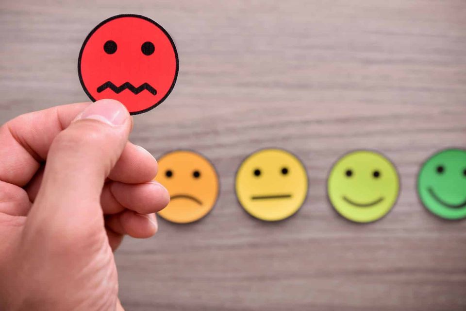 A red card with a sad face is held up by a hand, with four other circular cards on a wooden surface in orange, yellow, light green, and green. These cards display facial expressions ranging from sad to happy.