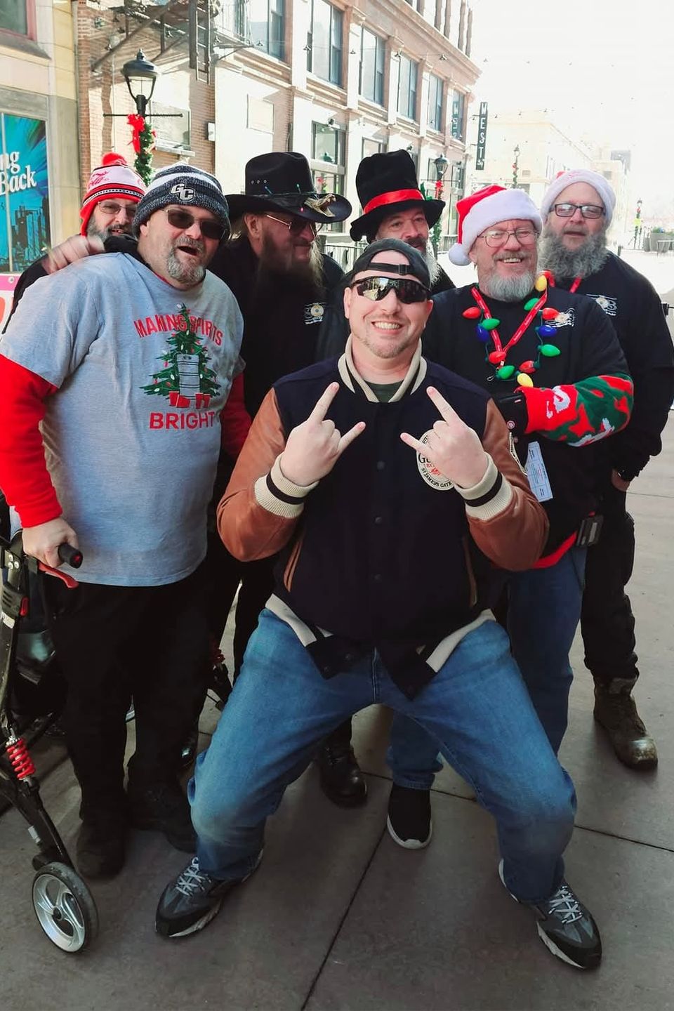 Group of attendees wearing Christmas hats, sweaters, and holiday accessories posing for a photo on a city sidewalk.
