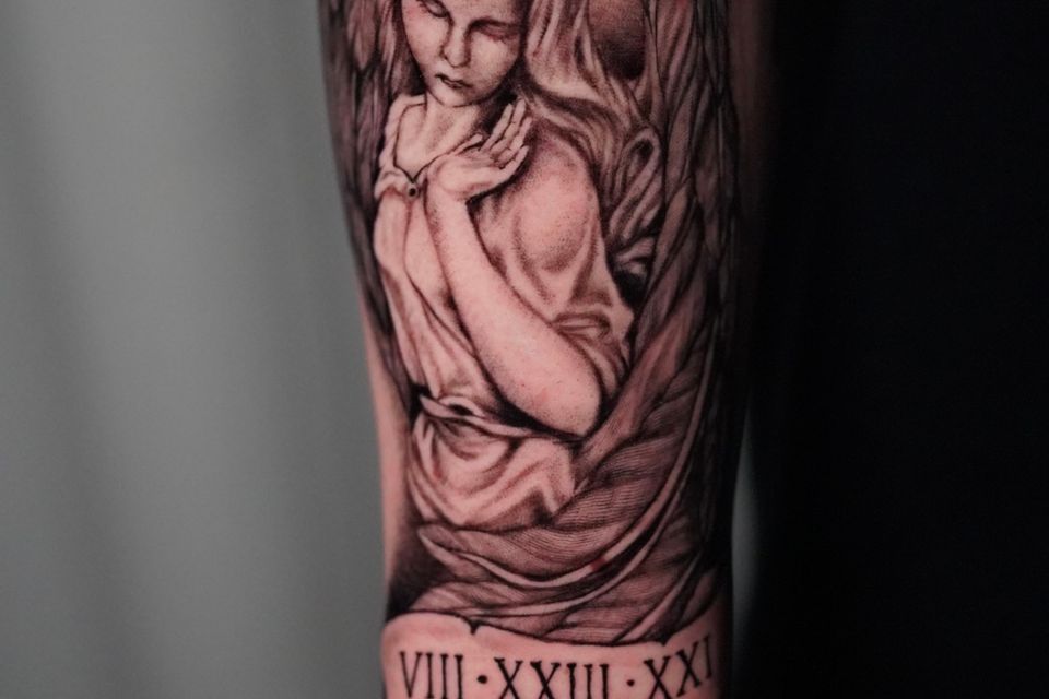 Image of a man's forearm with an angel tatto and Roman numerals below in black and grey ink