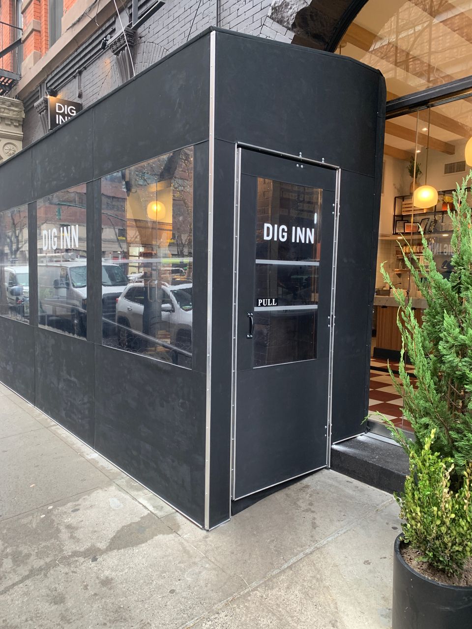 Black seasonal vestibule with clear panels and a door reading Dig Inn built out onto the sidewalk in front of a restaurant
