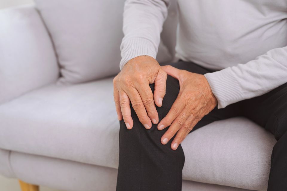 Senior man sitting on sofa in living room at home and touching his knee by the pain