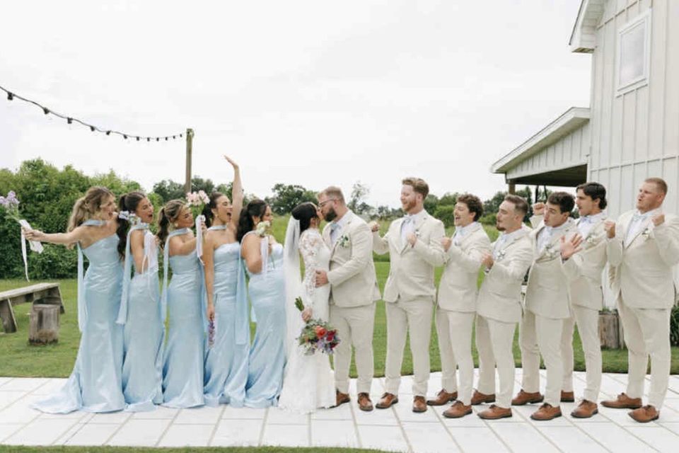 Bride and groom kissing outdoors while bridesmaids in light blue dresses and groomsmen in beige suits cheer around them.