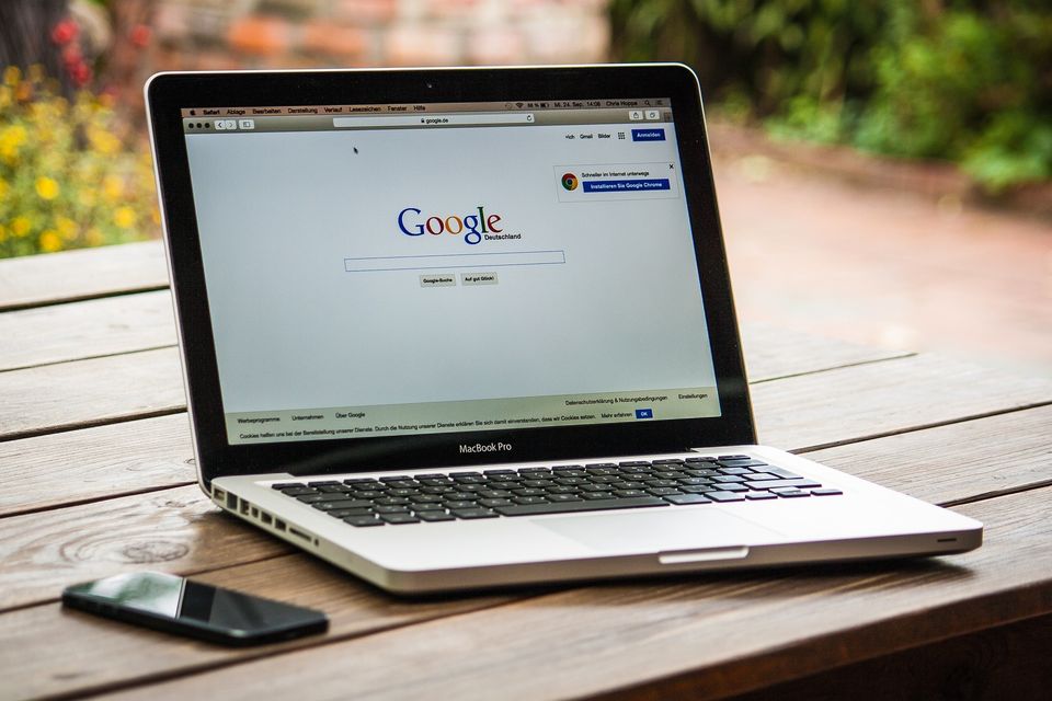 Google search screen on a MacBook Pro laptop sitting on a table with a black cell phone.