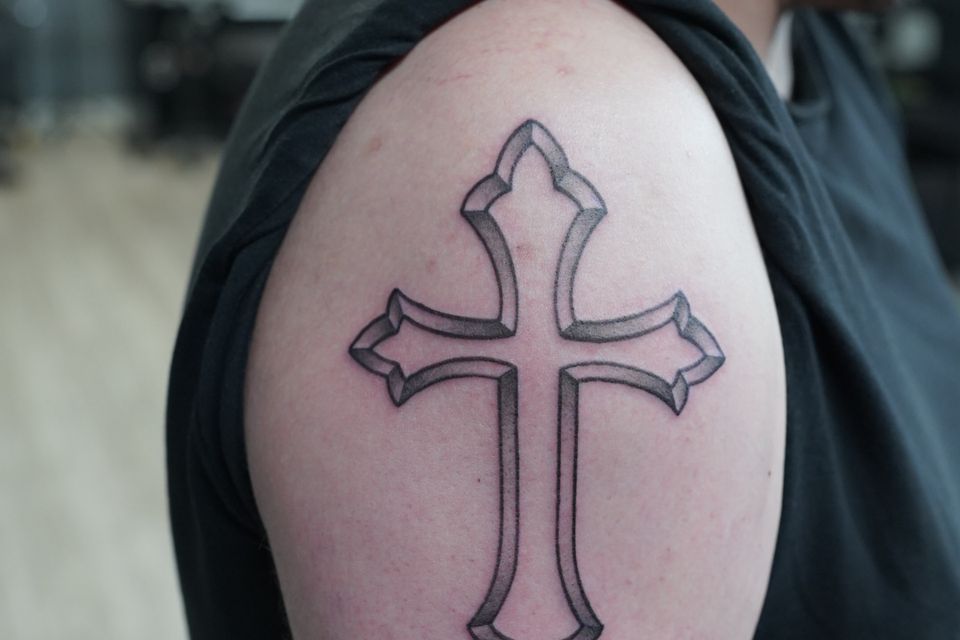 Image of a man's right shoulder bearing a tattoo of an ancient cross in black, grey and white ink