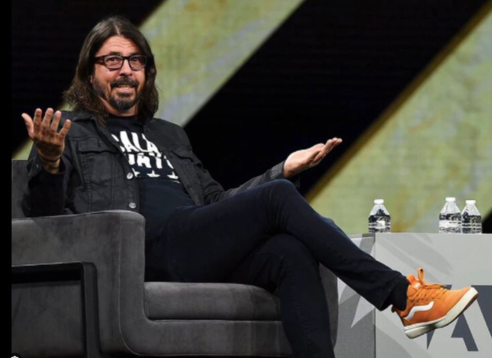 Dave Grohl in his orange vans sitting on a chair in black jeans, a black t-shirt and black flannel- wearing his glasses. 