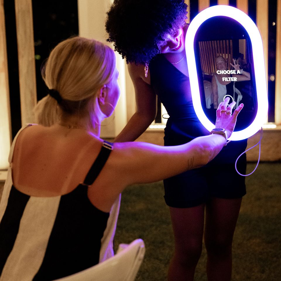 Two women using a roaming photo booth screen to select a filter at an outdoor evening event.