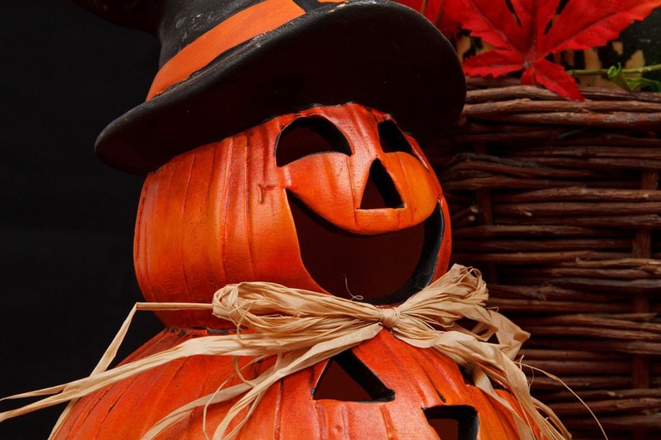 Stacked jack-o'-lantern pumpkin decoration with carved faces, topped with a black witchâs hat and raffia bow, surrounded by a wicker basket, patterned gourd, and red autumn leaves, set against a dark background. Halloween 21498 1280