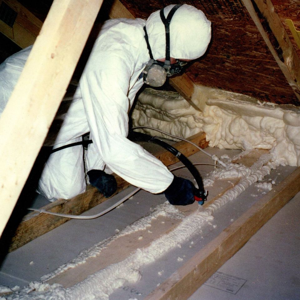 Technician applying spray foam insulation inside an attic while wearing protective clothing and a respirator.
