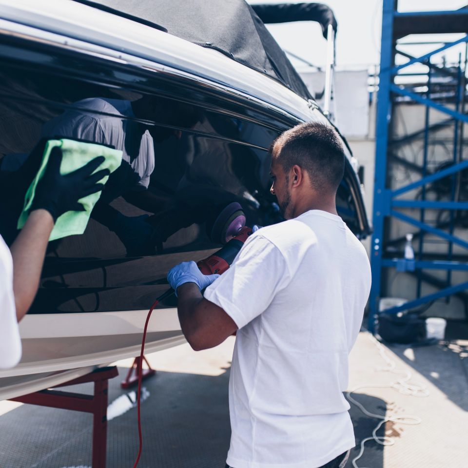 Two technicians polishing and wiping down a dark boat hull on stands during marine and boat detailing