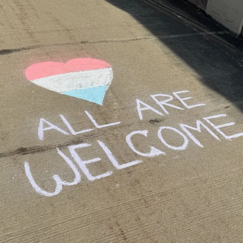 Chalk on sidewalk with a 3 color heart in pink, white and light blue and the words "all are welcome"
