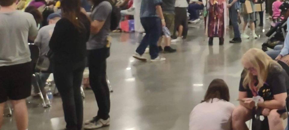 Crowd of attendees walking, chatting, and seated inside a busy comic convention hall, with people dressed in casual and cosplay outfits enjoying the lively atmosphere.