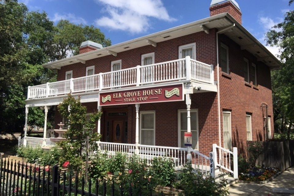 Historic Elk Grove House Stage Stop with red brick facade, white trim, wraparound porch and balcony, and garden landscaping under clear skies