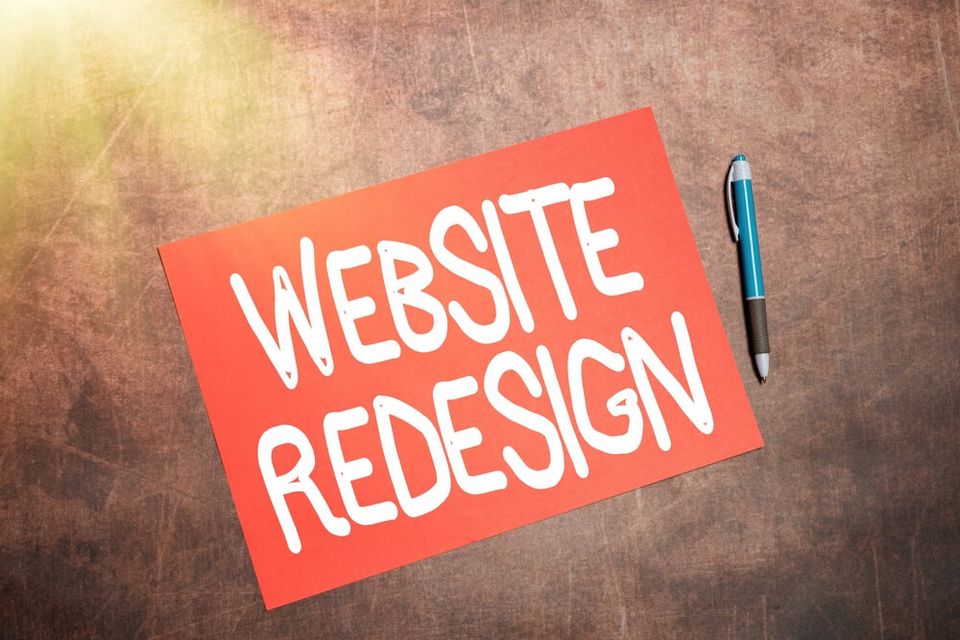 Red card featuring the bold, white text 'WEBSITE REDESIGN,' placed on a rustic wooden surface with a black and blue pen lying next to it.
