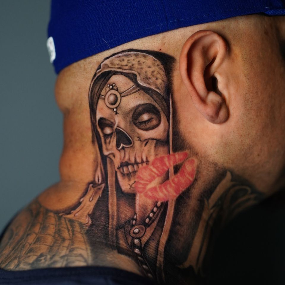 Image of a skeleton tattoo dressed up like a princess and located on the clients neck extending to about the middle of his ear, the tattoo is in black and grey except for a pair of red lips superimposed over the main tattoo.