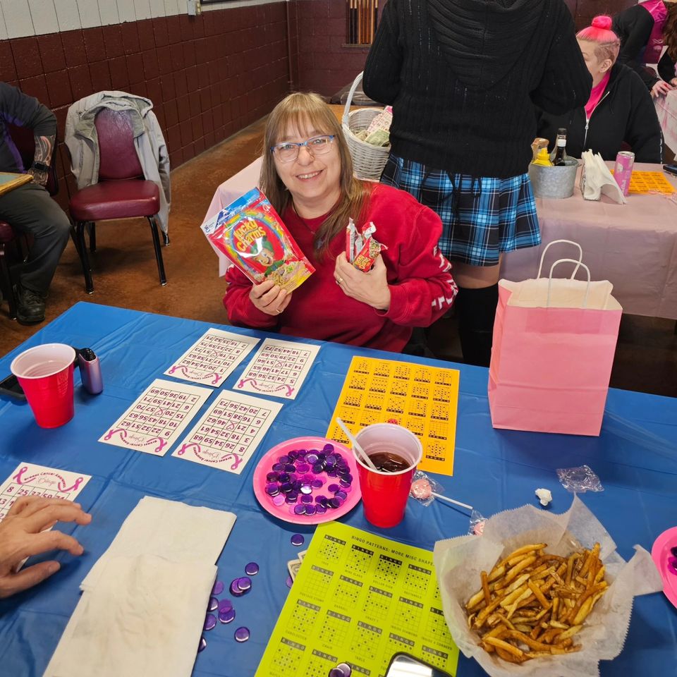 Smiling bingo player holding prize items at a decorated bingo table with cards, game markers, drinks, and food.