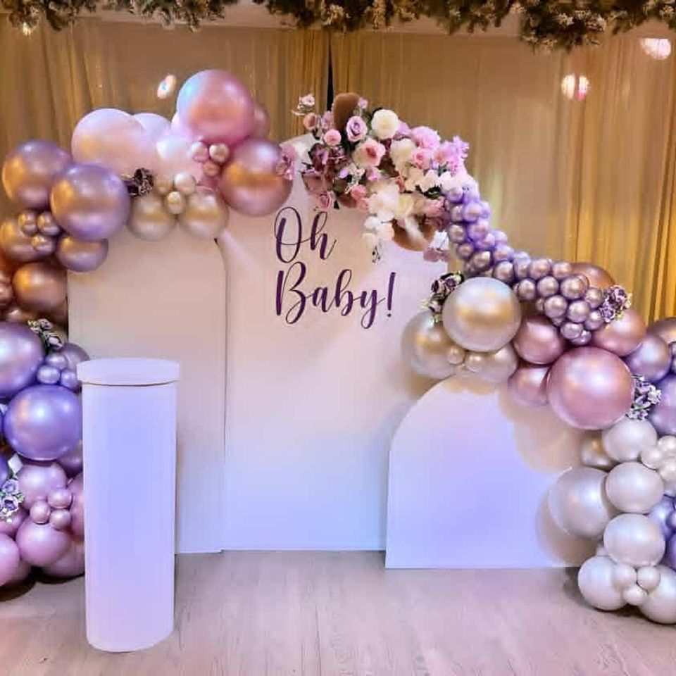 Elegant baby shower backdrop with metallic pink, purple, and silver balloon garland, floral accents, and ‘Oh Baby!’ sign.