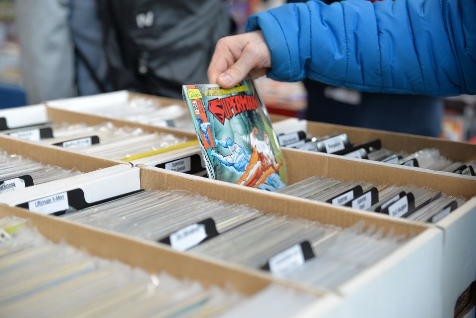 Comic book shop/vendor.  Person wearing a blue jacket picking up a vintage Spider-Man comic book from a long box filled with neatly organized, bagged, and boarded comics at a convention or vendor table.