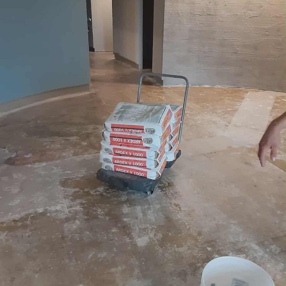 Concrete floor with bags of flooring material on a cart and a bucket in an unfinished interior space.