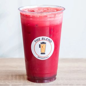 A deep red-purple juice in a clear "The Blend Smoothies" cup, representing a mix of beet, carrot, apple, ginger, and lemon on a wooden surface.
