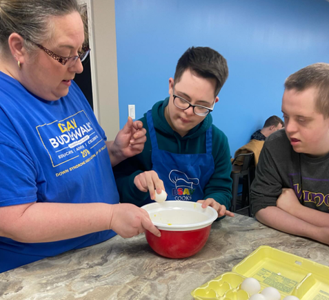 Instructor helping participants with Down syndrome crack eggs into a mixing bowl during a cooking class.