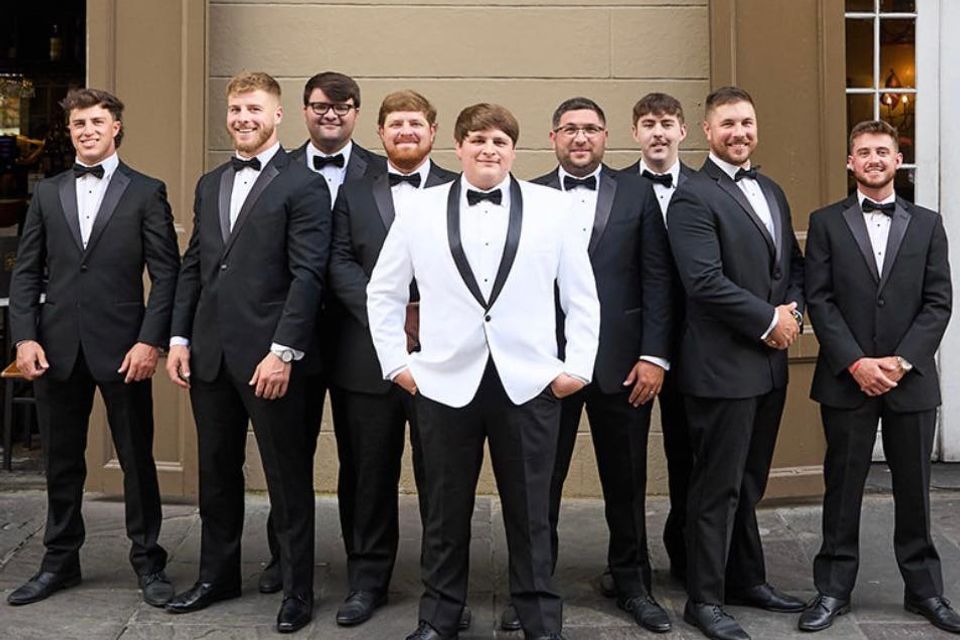 Groom and groomsmen wearing classic black tuxedos, with the groom in a white tuxedo jacket, posing outdoors.
