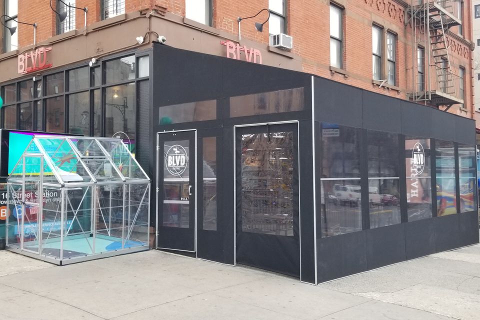 Long black framed seasonal vestibule running along a storefront and wrapping the corner, with a clear paneled enclosure beside it on the sidewalk