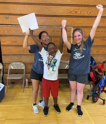 A child raises both arms in celebration alongside two volunteer coaches after completing a biking milestone at DSAV's iCan Shine Bike Camp.