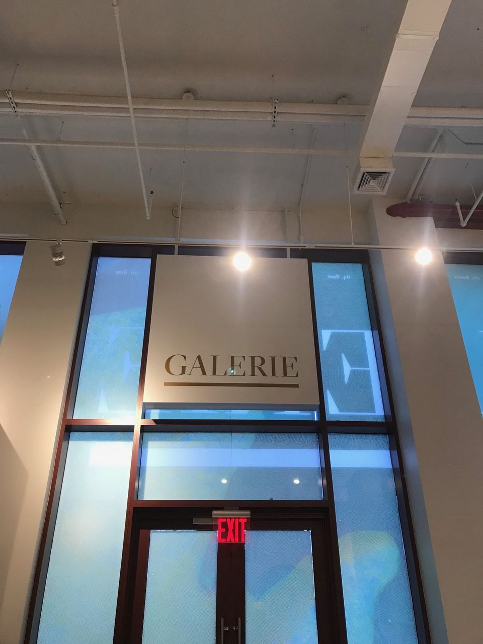 Illuminated panel reading Galerie mounted above tall glass entry doors inside a building lobby