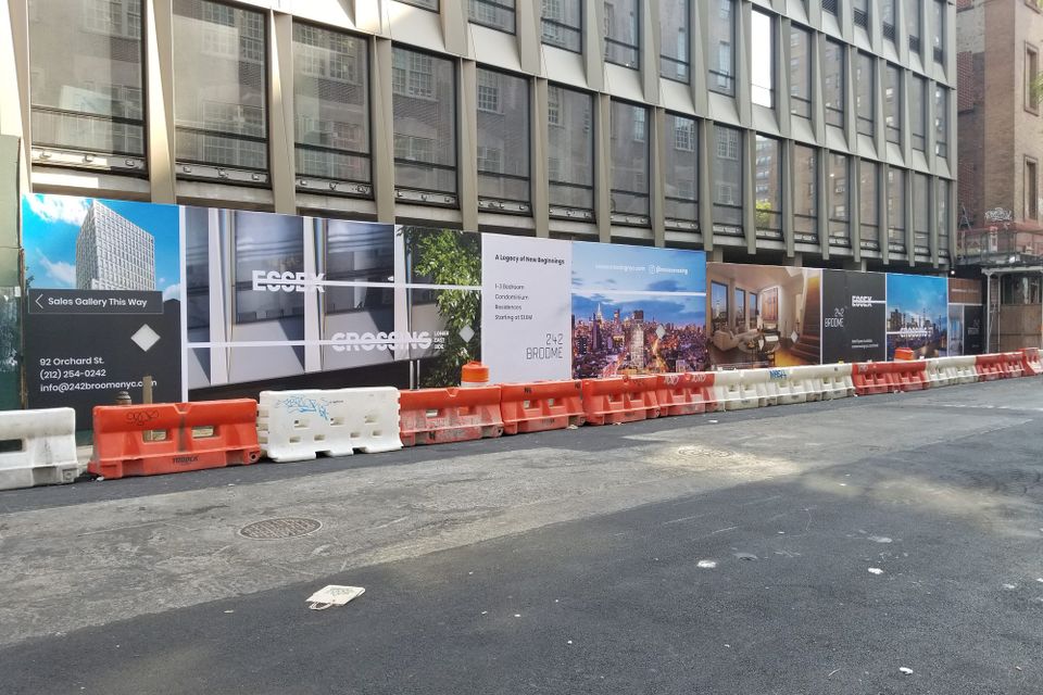 Printed construction hoarding graphics for 242 Broome running along a sidewalk behind a row of traffic barriers