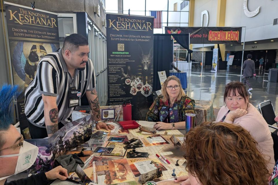 David Dean Haddon running a tabletop RPG session at GameHoleCon with banners for The Kingdom of Keshanar and players seated around a game table