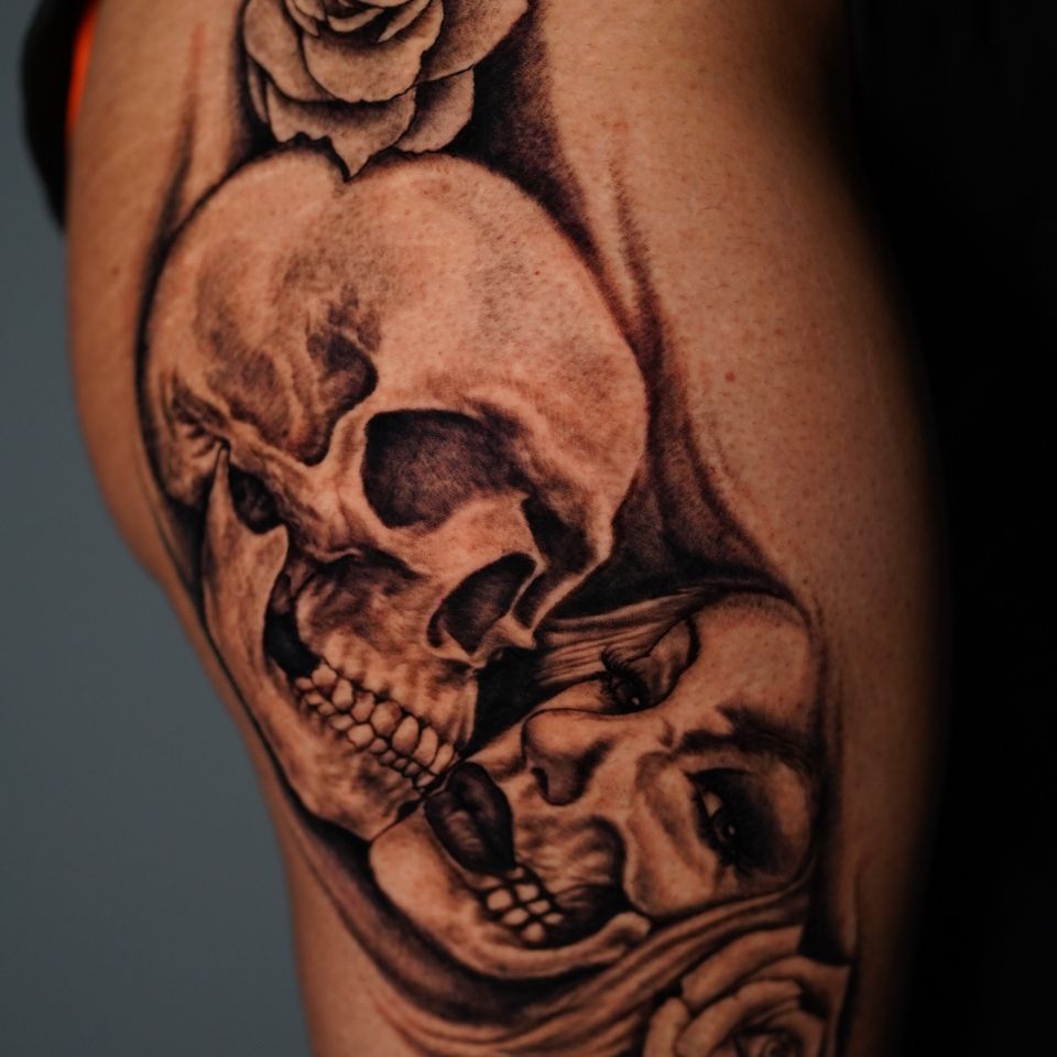 Image of an attractive ish woman who is tattood in black and has partial skull showing next to a full skull that appears to be larger, maybe a male, maybe a husban who passed away, there are two flowers one on each side of the tattoo