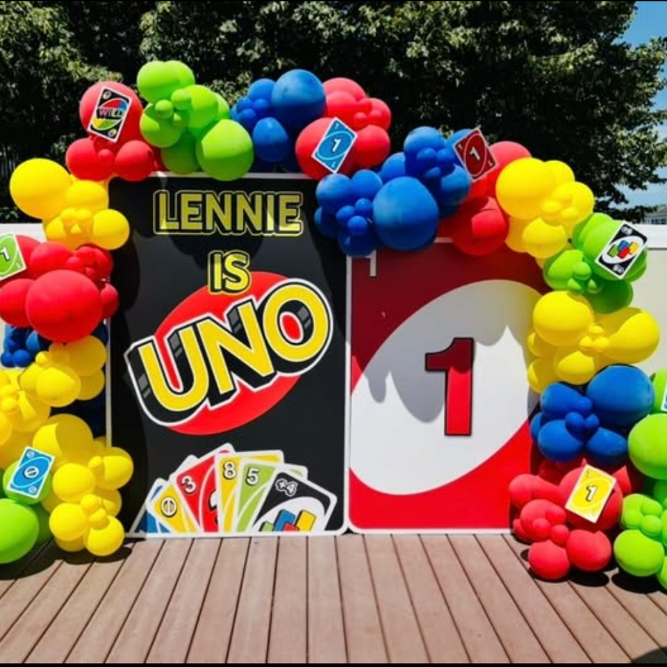 UNO themed birthday party backdrop with colorful red, yellow, green, and blue balloon garland, giant UNO cards, and custom name display for Lennie.